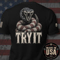 Black short-sleeve t-shirt shown from the back with a large graphic of a coiled snake above distressed white text reading 'TRY IT'. The shirt is photographed against a rustic American flag background, with a circular 'Made in USA 100% American Made' badge overlaid in the lower right corner.