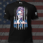 A black short-sleeve graphic T-shirt displayed against a distressed American flag background. The shirt features a central graphic of a woman with purple hair wearing sunglasses with an American flag pattern, with 'MADE IN AMERICA' printed below in red and blue.