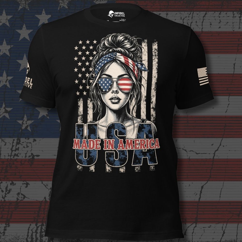 A black short-sleeve graphic T-shirt featuring a stylized American flag design with a woman's face wearing sunglasses, distressed vertical stripes, and the text 'USA MADE IN AMERICA' across the front. The shirt is shown against a rustic American flag backdrop.