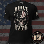 A black short-sleeve graphic T-shirt displayed against a distressed American flag background. The shirt features a large front graphic of a skull with an American flag pattern inside an oval, with the words 'BUILT FROM 1776' in distressed white lettering.