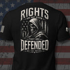 Black short-sleeve t-shirt shown from the back, featuring a large distressed white graphic of a hooded skull over an American flag with the text 'RIGHTS ARE NOT GIVEN THEY ARE DEFENDED WE THE INFIDEL'. The shirt is displayed against a rustic American flag backdrop.