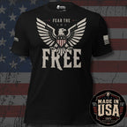 A black short-sleeve graphic T-shirt displayed against a distressed American flag background. The shirt features a large eagle graphic with outstretched wings and the words 'FEAR THE FREE' in distressed white and gray lettering. A round emblem indicates this design is 100% American Made - Made in the USA.