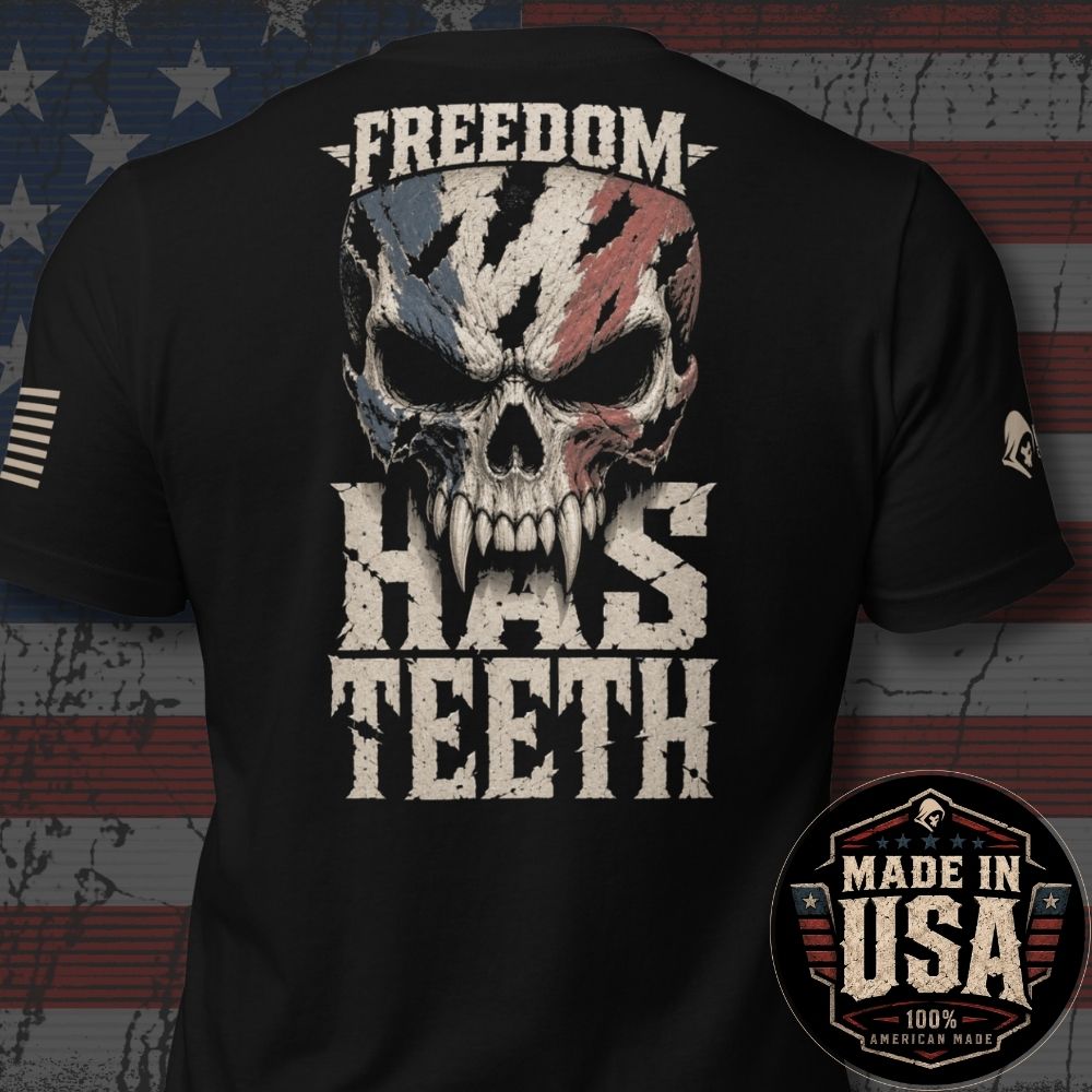 Black short-sleeve t-shirt shown from the back, featuring a large distressed graphic of a skull with an American flag pattern and the text 'FREEDOM HAS TEETH'. A circular 'MADE IN USA 100% American Made' badge is overlaid in the bottom right corner.