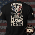 Black short-sleeve t-shirt shown from the back, featuring a large distressed graphic of a skull with an American flag pattern and the text 'FREEDOM HAS TEETH'. A circular 'MADE IN USA 100% American Made' badge is overlaid in the bottom right corner.