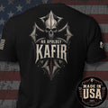 Black short-sleeve t-shirt shown from the back with a large distressed graphic featuring a skull, crossed elements, and the text 'NO APOLOGY KAFIR'. A circular 'MADE IN USA 100% AMERICAN MADE' badge appears in the lower right corner, and the shirt is photographed against a rustic American flag background.