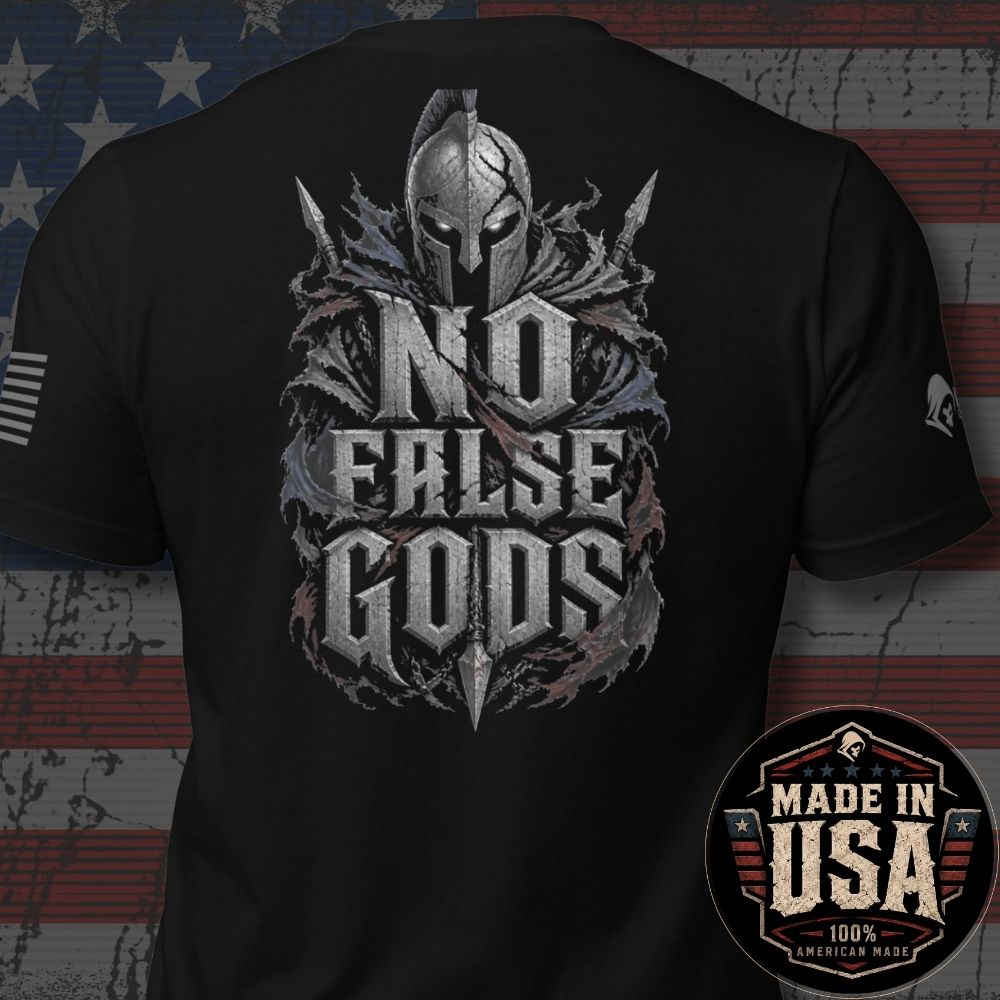 Black short-sleeve t-shirt shown from the back, featuring a large distressed graphic of a Spartan helmet and crossed swords with the text 'NO FALSE GODS' in metallic gray. The shirt is displayed against a rustic American flag background, with a circular 'Made in USA 100% American Made' badge in the bottom right corner.