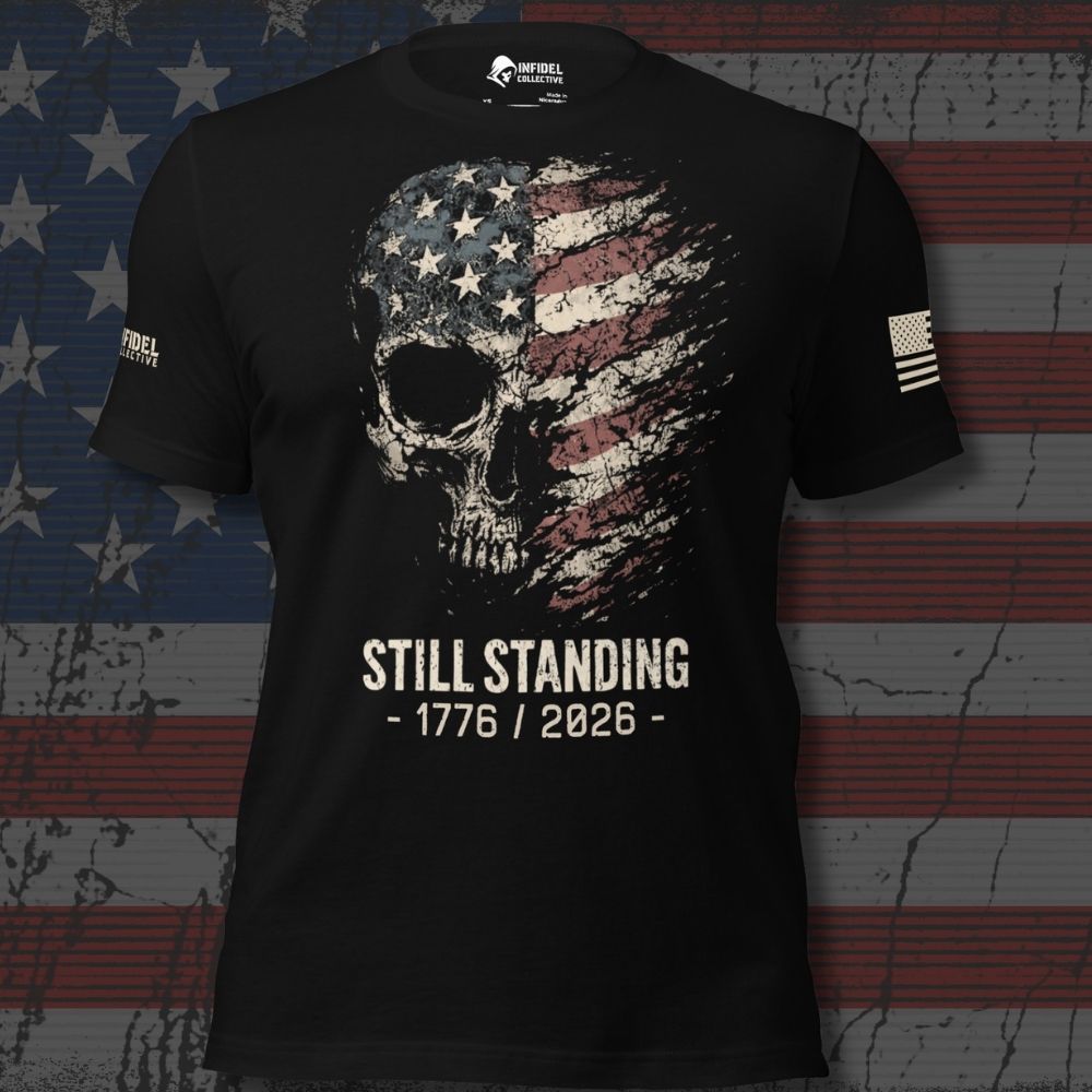 A black short-sleeve graphic T-shirt displayed flat against a distressed American flag background. The shirt features a large skull graphic filled with an American flag pattern, with the text 'STILL STANDING - 1776 / 2026 -' printed below in white.