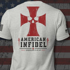 A light heather gray t-shirt shown from the back, featuring a large red Iron Cross with a skull design, the words 'AMERICAN INFIDEL' in black, and 'EPHESIANS 6:10-18' printed below. The shirt is displayed against a distressed American flag background.