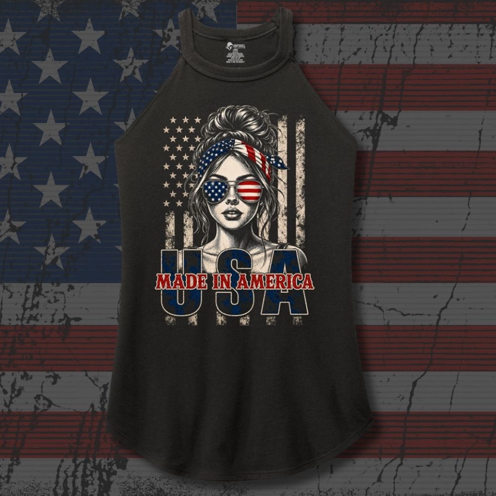 A black raglan sleeveless tank top featuring a patriotic graphic of a woman with an American flag headband and sunglasses, with the text 'MADE IN AMERICA' and 'USA' printed below, displayed against a distressed American flag background.