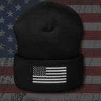 A black knit beanie with a folded cuff featuring a white embroidered American flag design, shown against a distressed American flag background.
