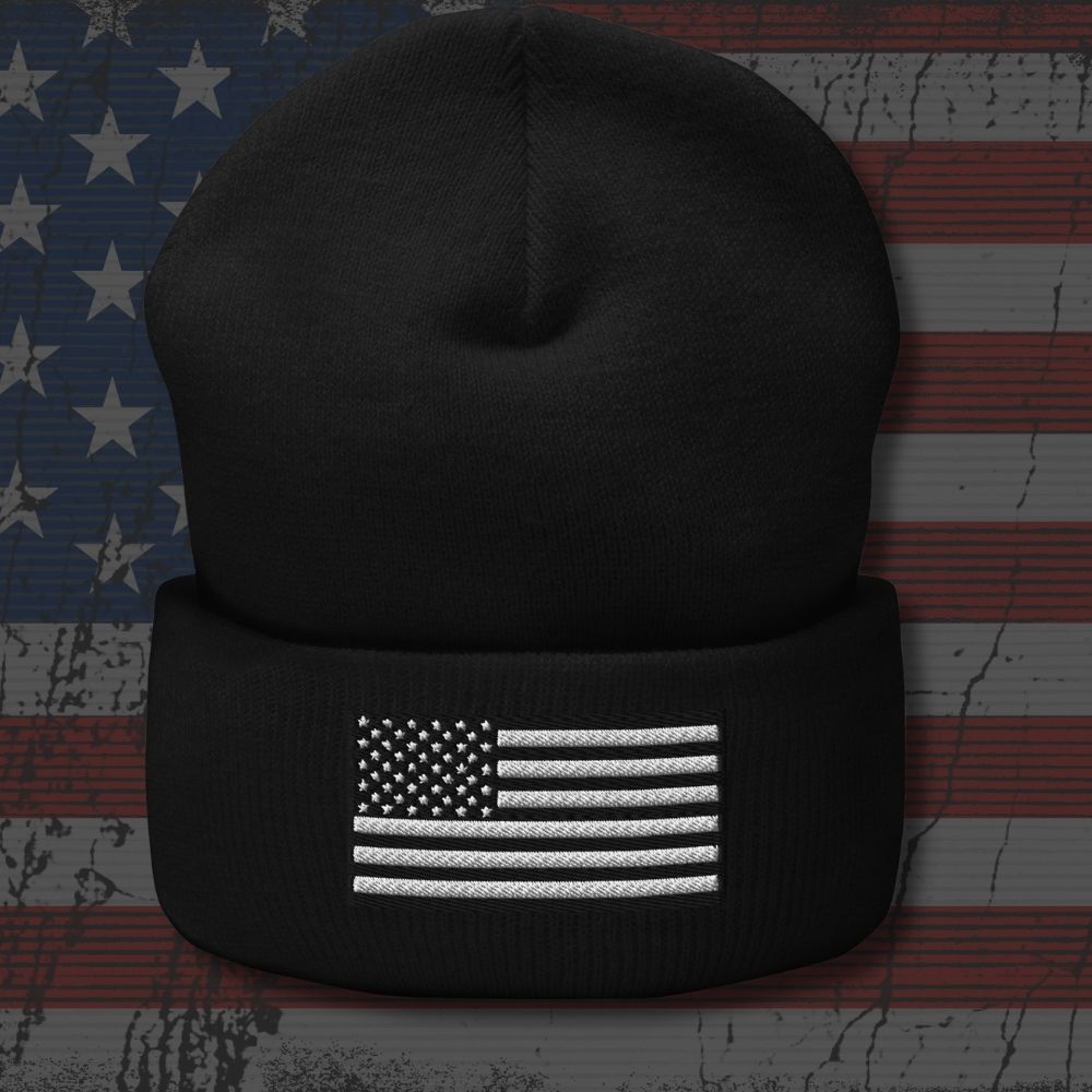 A black knit beanie with a folded cuff featuring a white embroidered American flag design, shown against a distressed American flag background.
