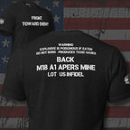 Claymore inspired design with "FRONT TOWARD ENEMY" across the front and "M18 A1 APERS MINE LOT IS-INFIDEL" across the black on a black tee against a USA flag background.