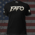 A black short-sleeve t-shirt with a large distressed white 'FAFO' graphic across the chest, featuring a power symbol inside the 'O' and additional white text on the right sleeve. The shirt is displayed against a distressed American flag background.