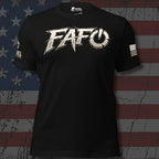 A black short-sleeve t-shirt with a large distressed white 'FAFO' graphic across the chest, featuring a power symbol inside the 'O' and additional white text on the right sleeve. The shirt is displayed against a distressed American flag background.
