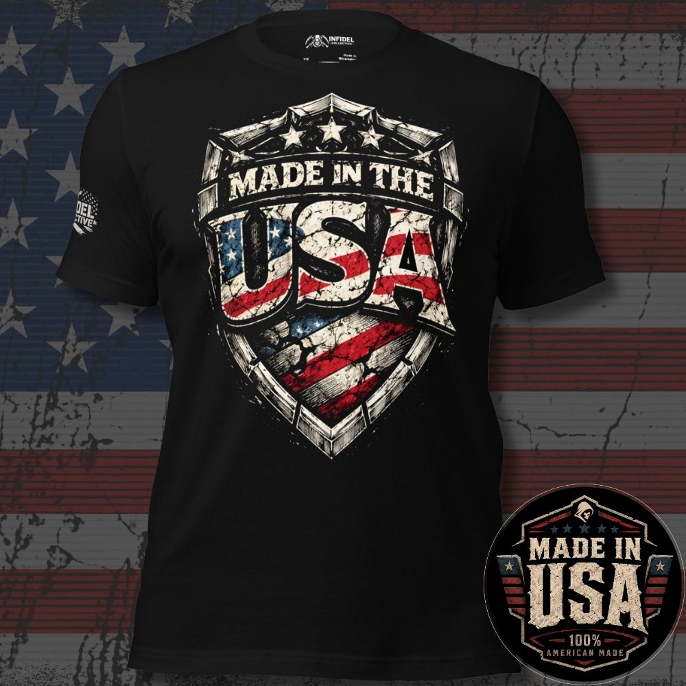 A black short-sleeve graphic T-shirt displayed against a distressed American flag background. The shirt features a large shield-style chest graphic reading "MADE IN THE USA" with an American flag pattern inside the letters.